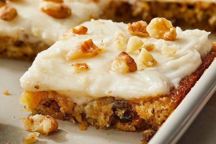 A close-up of a frosted bar topped with chopped nuts, featuring a moist, golden-brown cake base. The frosting is creamy and white, and the dessert is served in a rectangular pan.