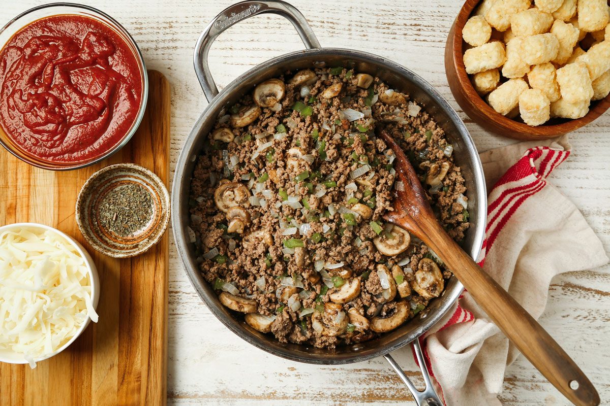 Overhead shot of a large skillet cook the beef; green pepper onion and mushrooms over medium heat until meat is no longer pink; breaking meat into crumbles; drain; add pizza sauce and basil; wooden board; wooden spoon; napkin; all set on a wooden surface;