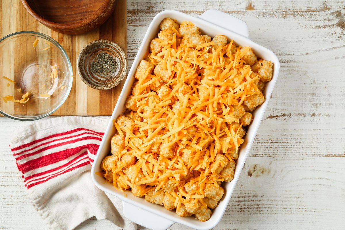 Overhead shot of Pizza Tater Tot Casserole; in a baking dish; sprinkle with cheddar cheese; bake until cheese is melted about 5 minutes; a napkin is nearby; all set on a wooden surface;