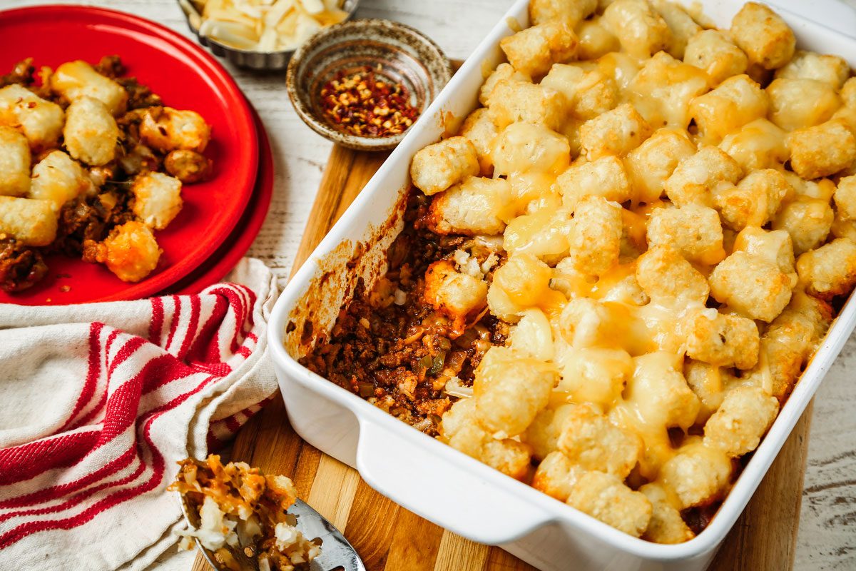 Overhead shot of Pizza Tater Tot Casserole; in a baking dish over a wooden board; served on red plate; a serving spoon and a napkin are nearby; all set on a wooden surface;