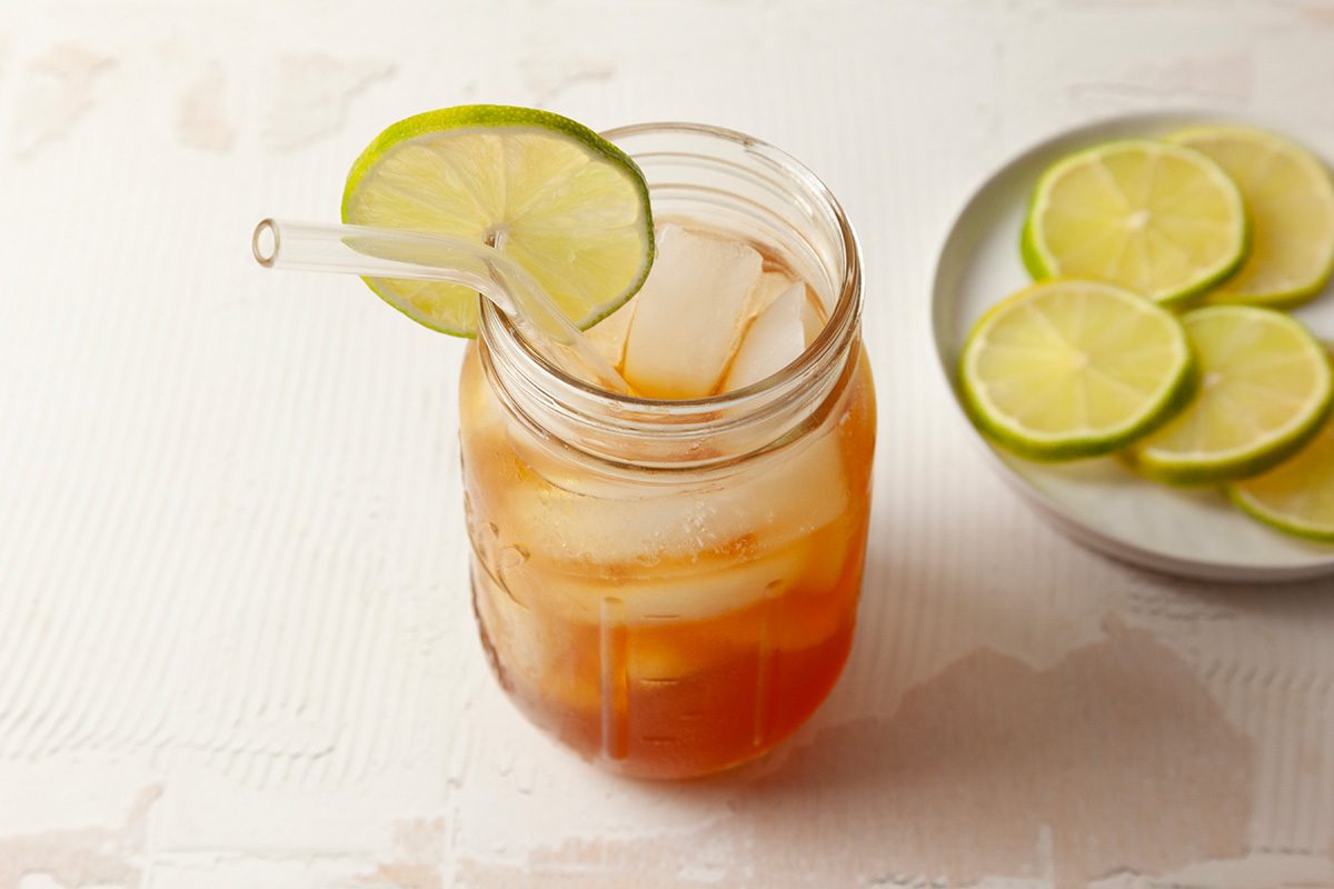 A mason jar filled with iced tea, ice cubes, and a lime slice garnish, with a clear straw. In the background, a plate holds several slices of lime on a light, textured surface.