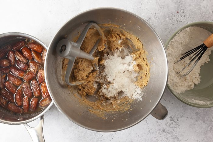 Three bowls on a countertop: one with soaked dates in liquid, one with partially mixed cookie dough and flour in a mixer bowl, and one with flour being whisked.