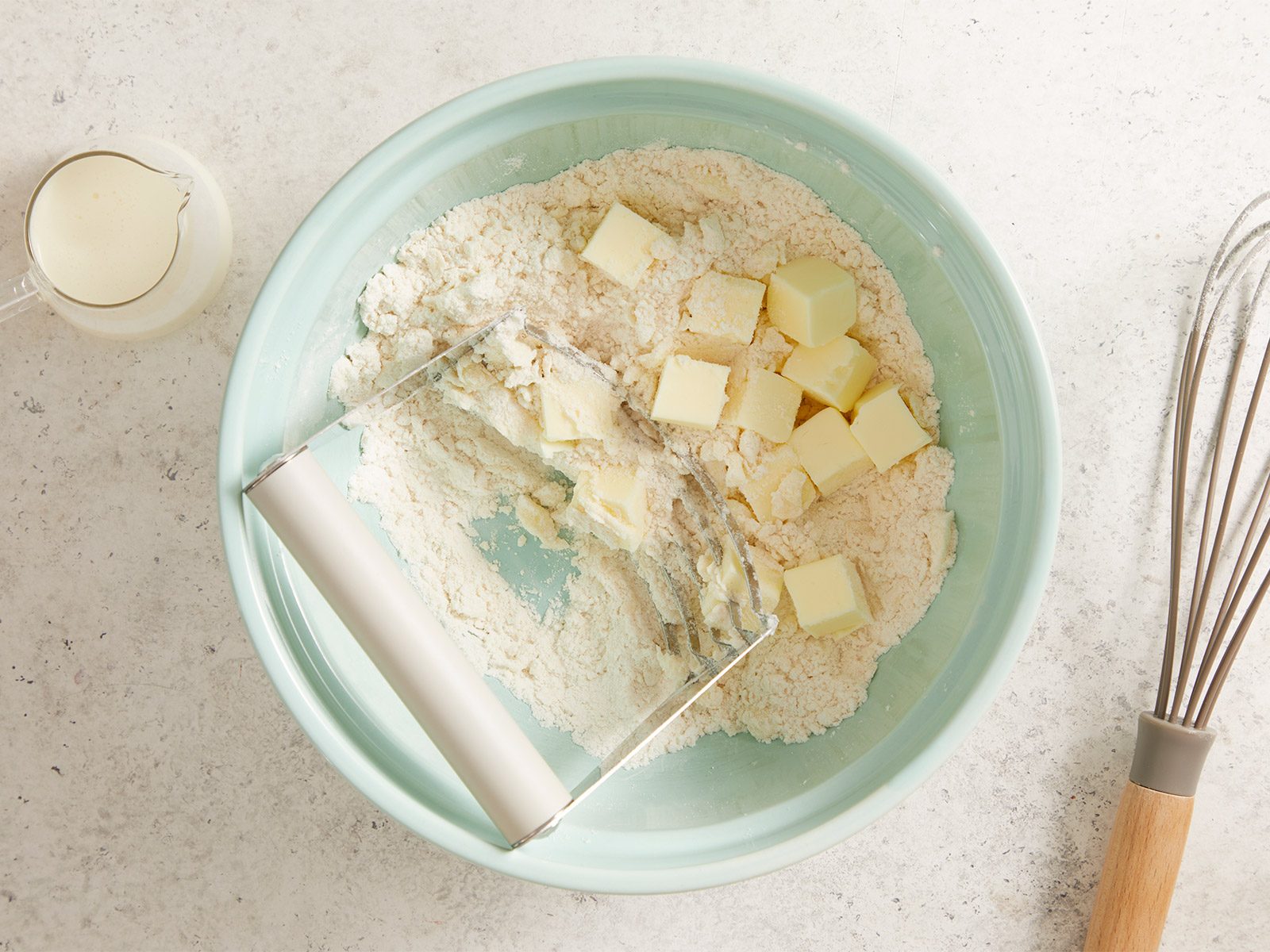 A light blue bowl holds flour and cubes of butter being mixed with a pastry blender. A glass measuring cup with milk and a whisk are nearby on a light countertop.