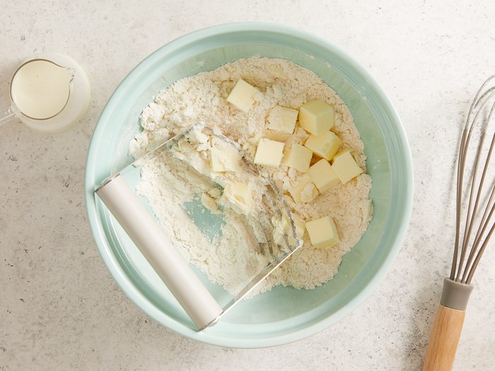 A light blue bowl holds flour and cubes of butter being mixed with a pastry blender. A glass measuring cup with milk and a whisk are nearby on a light countertop.