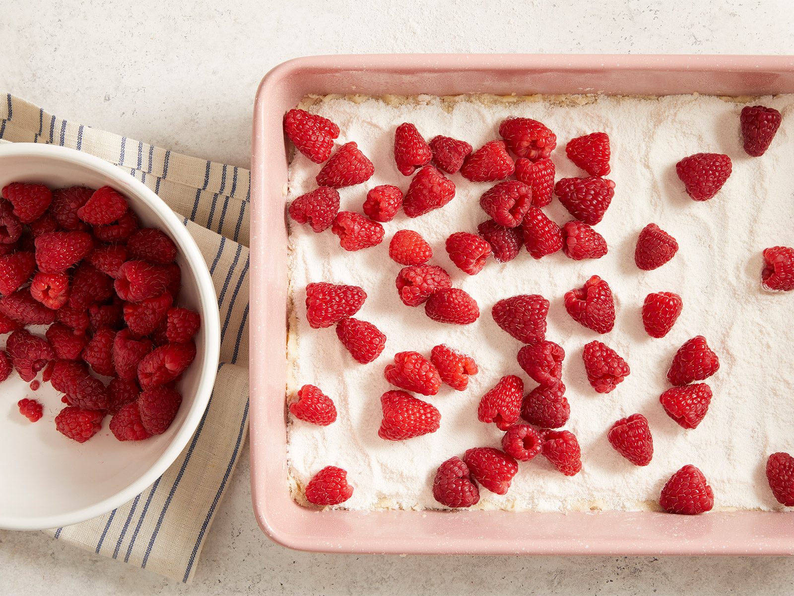 A pink baking dish filled with a dessert topped with powdered sugar and fresh raspberries sits next to a white bowl of loose raspberries on a striped cloth.