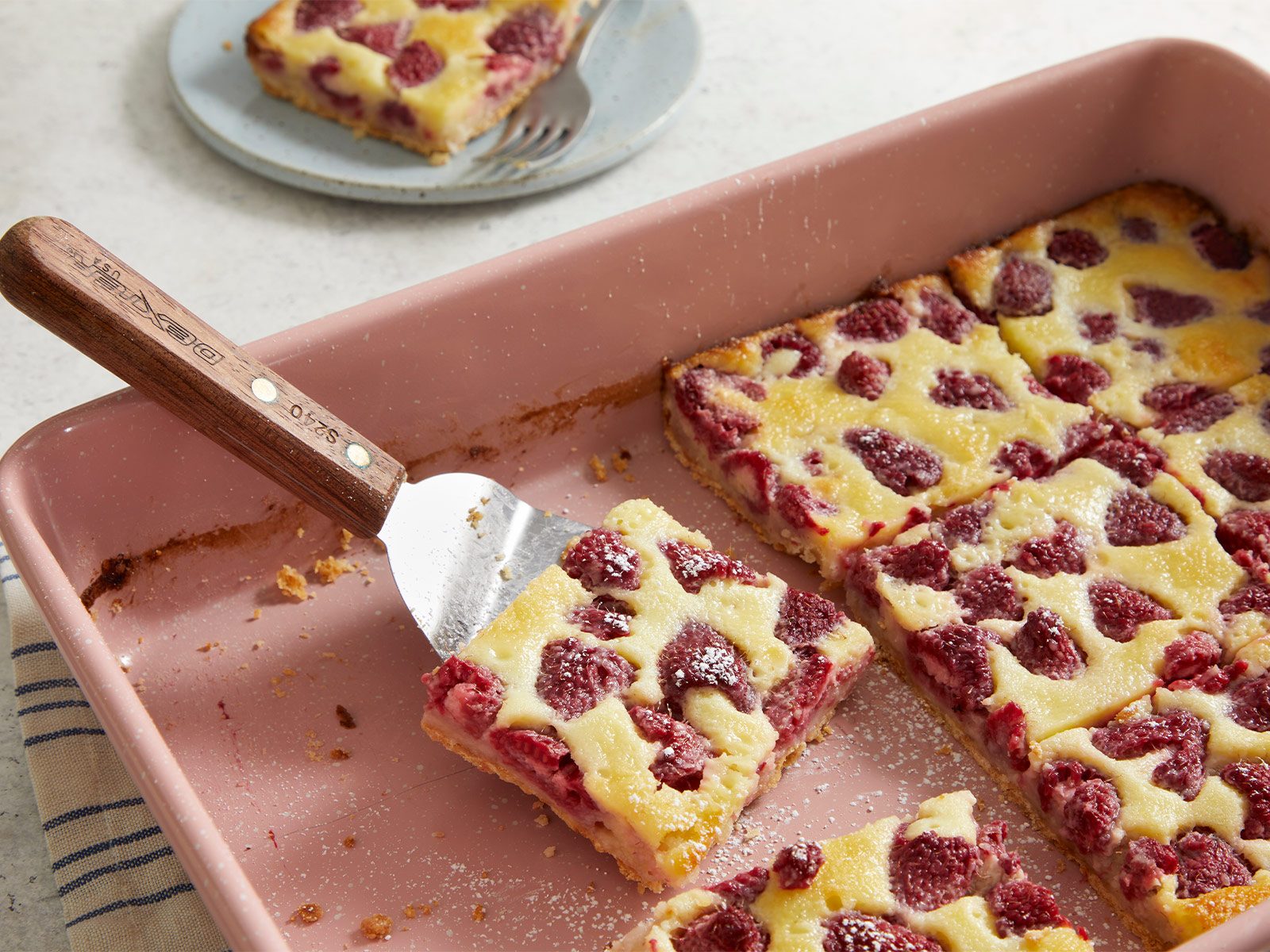 A baking dish with raspberry dessert bars, some cut into squares. A serving spatula lifts one bar, showing a golden crust, creamy filling, and fresh raspberries. A plate with another bar sits in the background.