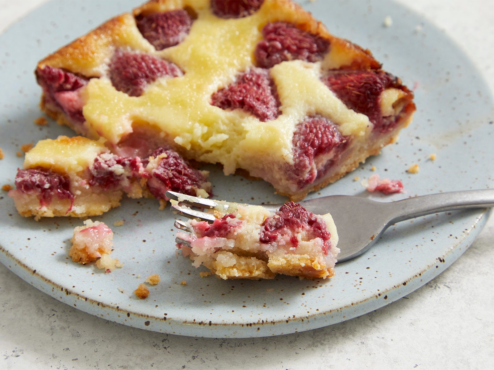 A square slice of raspberry dessert bar with a creamy topping sits on a speckled plate. A fork holds a bite-sized piece, showing layers of crust, creamy filling, and raspberries. Crumbs are scattered on the plate.
