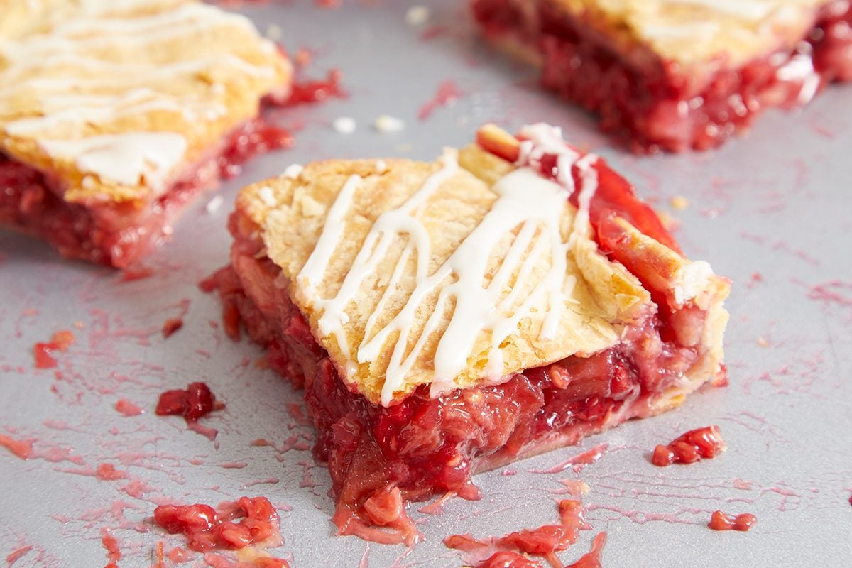 A close-up of a square slice of strawberry-filled pastry with a golden, flaky crust, drizzled with white icing. Pieces of the pastry and strawberry filling are visible on the surface around it.