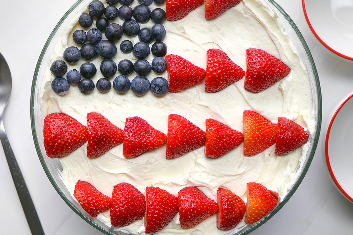 A dessert topped with whipped cream, blueberries arranged in a square, and rows of sliced strawberries, resembling the American flag. A spoon and two plates are nearby.