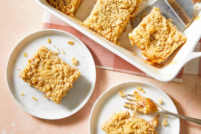 A baking dish with rectangular pieces of crumb-topped dessert, likely coffee cake or crumb bars. Two pieces are served on white plates, one with a fork, on a light surface with a checkered cloth underneath.