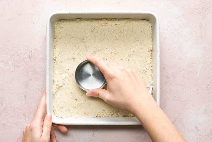 A hand presses a round metal cutter into a square pan filled with dough, on a light pink surface.