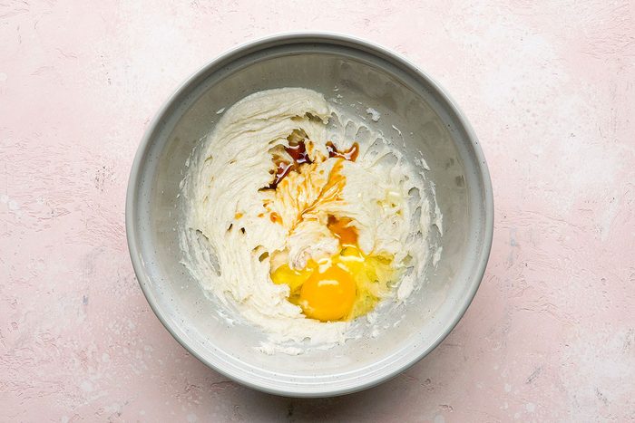 A gray bowl on a light pink surface contains cream cheese, an egg, vanilla extract, and some brown sugar or spices, partially mixed together.