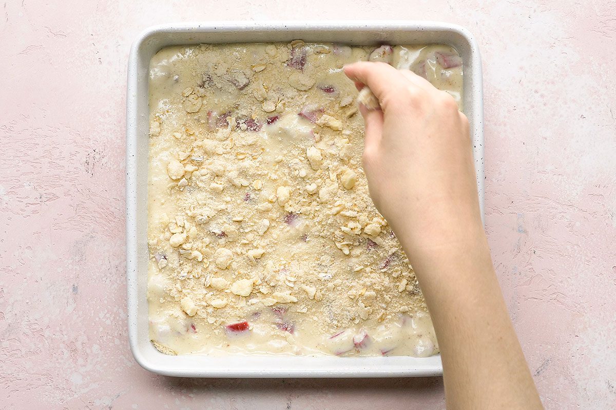 A hand sprinkles oat topping over an unbaked cake or dessert batter in a square baking dish on a light pink surface.