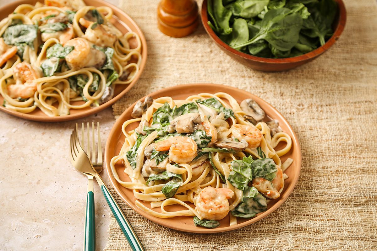 A plate of creamy pasta with shrimp, spinach, and mushrooms sits on a beige textured surface, with a fork and knife beside it. Another plate and a bowl of fresh spinach are in the background.