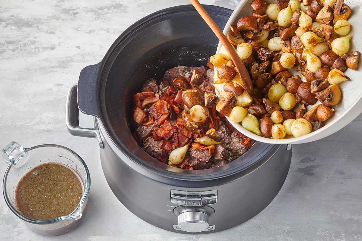 A bowl of cooked onions and mushrooms is being poured into a slow cooker filled with meat and bacon. A glass measuring cup with broth sits beside the slow cooker on a light-colored surface.