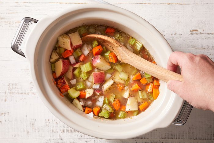 A hand stirs chopped vegetables, including potatoes, carrots, celery, and onions, in broth inside a slow cooker with a wooden spoon on a white wooden surface.