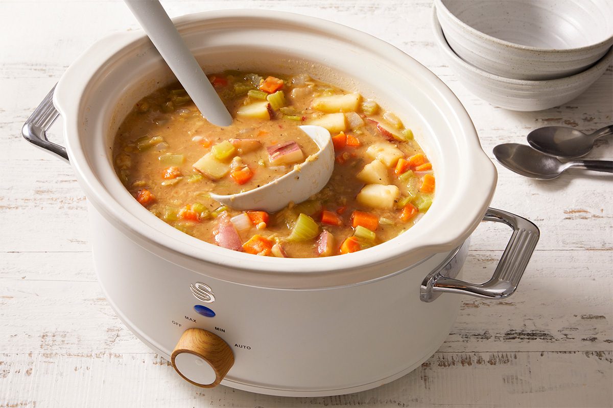A white slow cooker filled with chunky vegetable stew, including carrots, potatoes, and celery, sits on a white wooden surface. A ladle rests inside the cooker, and stacked bowls and spoons are in the background.