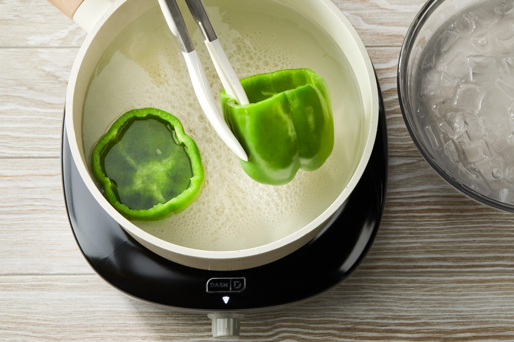Overhead shot of a skillet over a induction; blanch peppers in boiling water for 5 minutes; Drain and rinse in cold water; set aside; tongs; ice water glass bowl; all set on a cream-colored wooden surface;
