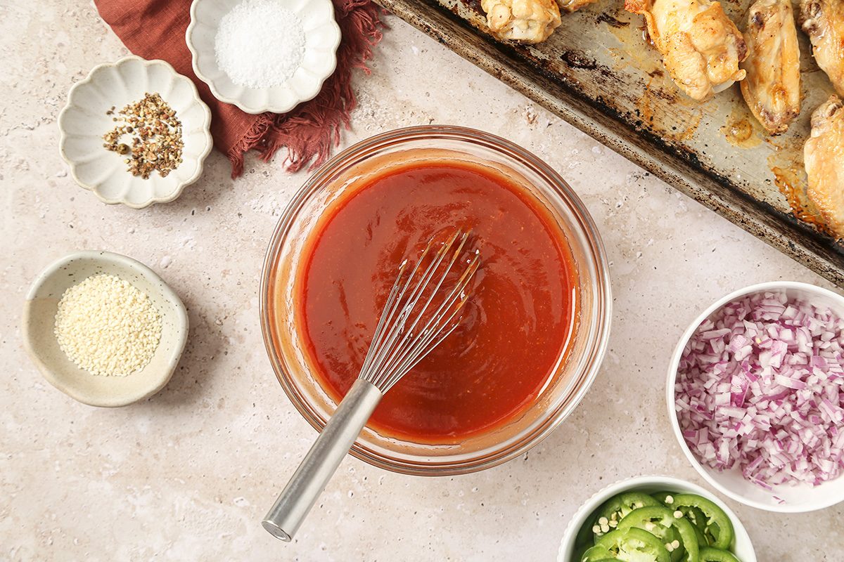 overhead shot of a glass bowl of red sauce with a whisk sits on a countertop, surrounded by bowls of sesame seeds, sliced jalapeños, chopped red onions, salt, pepper, and a tray of cooked chicken wings