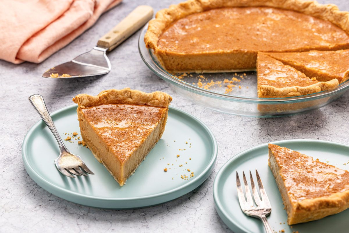 3/4 angle view shot of Apple Butter Pie in a glass pie plate; cut slices and served on two plates with forks; knife and a napkin resting nearby; all arranged on a grey marble surface;