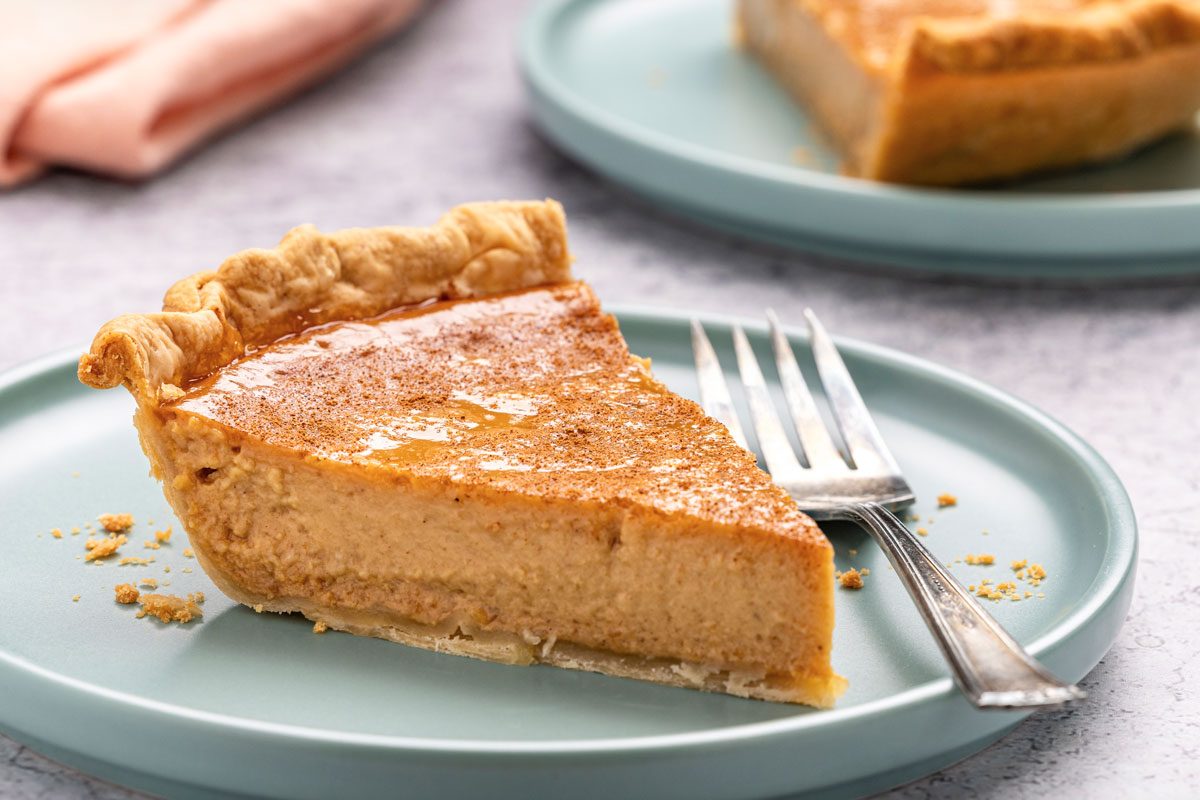 Closeup shot of Apple Butter Pie in a glass pie plate; cut slices and served on plate with fork; a napkin nearby; all arranged on a grey marble surface;