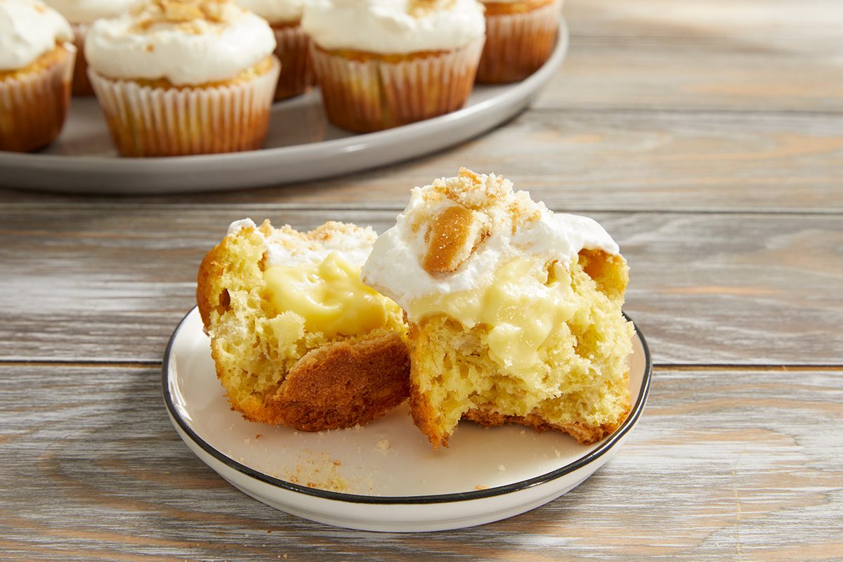A cupcake filled with creamy custard and topped with whipped frosting sits halved on a plate, with more frosted cupcakes in the background on a platter.
