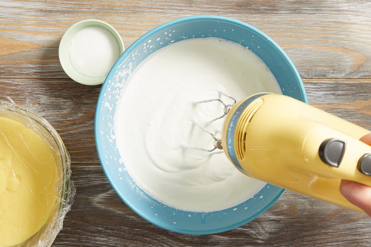 A hand is using a yellow electric mixer to beat egg whites in a blue bowl. Nearby, there is a bowl of thick yellow batter and a small bowl of white granulated sugar on a wooden surface.