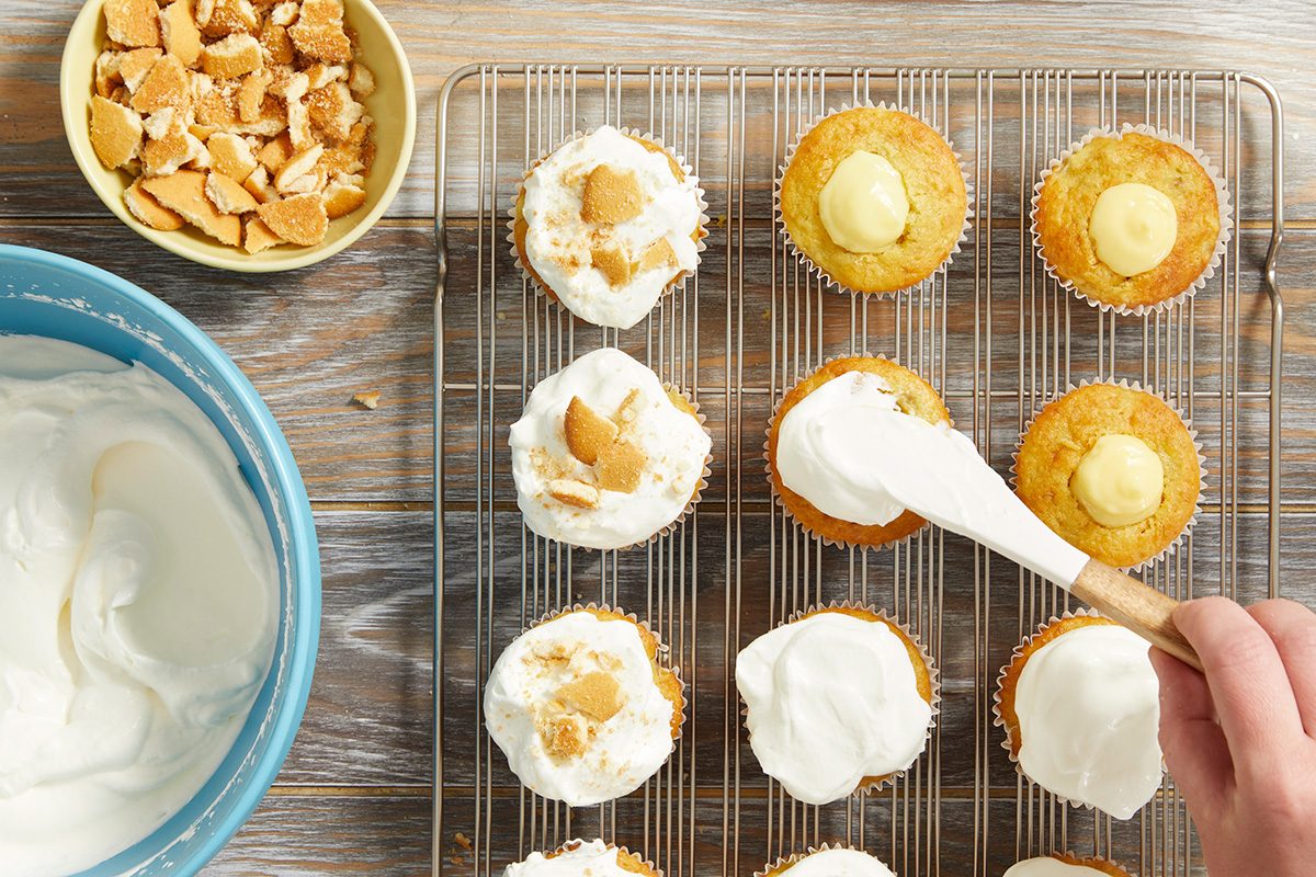 A hand spreads whipped cream onto cupcakes on a cooling rack. Some cupcakes are already topped with whipped cream and crumbled cookies. A bowl of whipped cream and a bowl of cookie pieces are nearby on a wooden surface.
