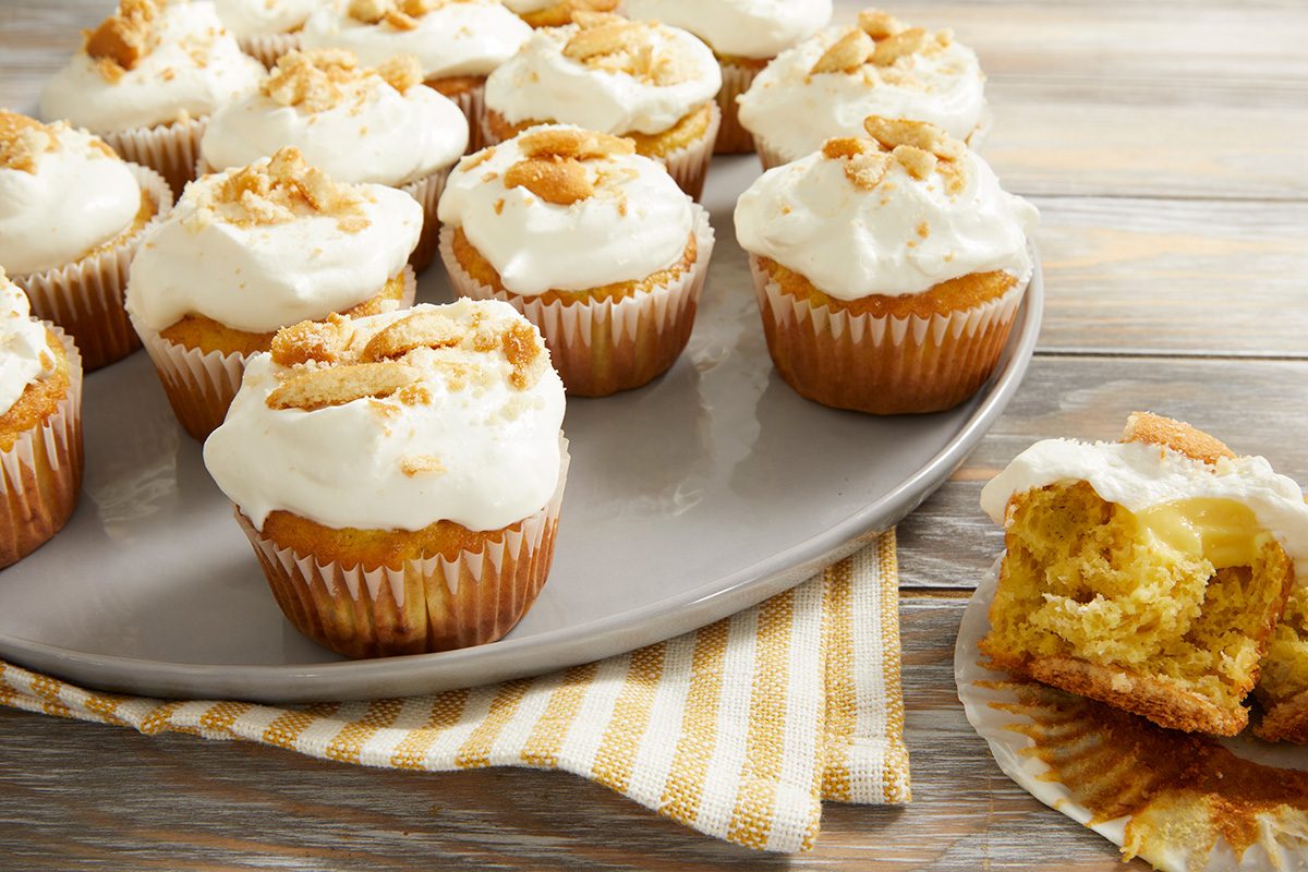 Cupcakes topped with white frosting and crumbled cookies are arranged on a gray plate. One cupcake is partially unwrapped with a bite taken out, showing a yellow, moist interior. The plate rests on a striped cloth.