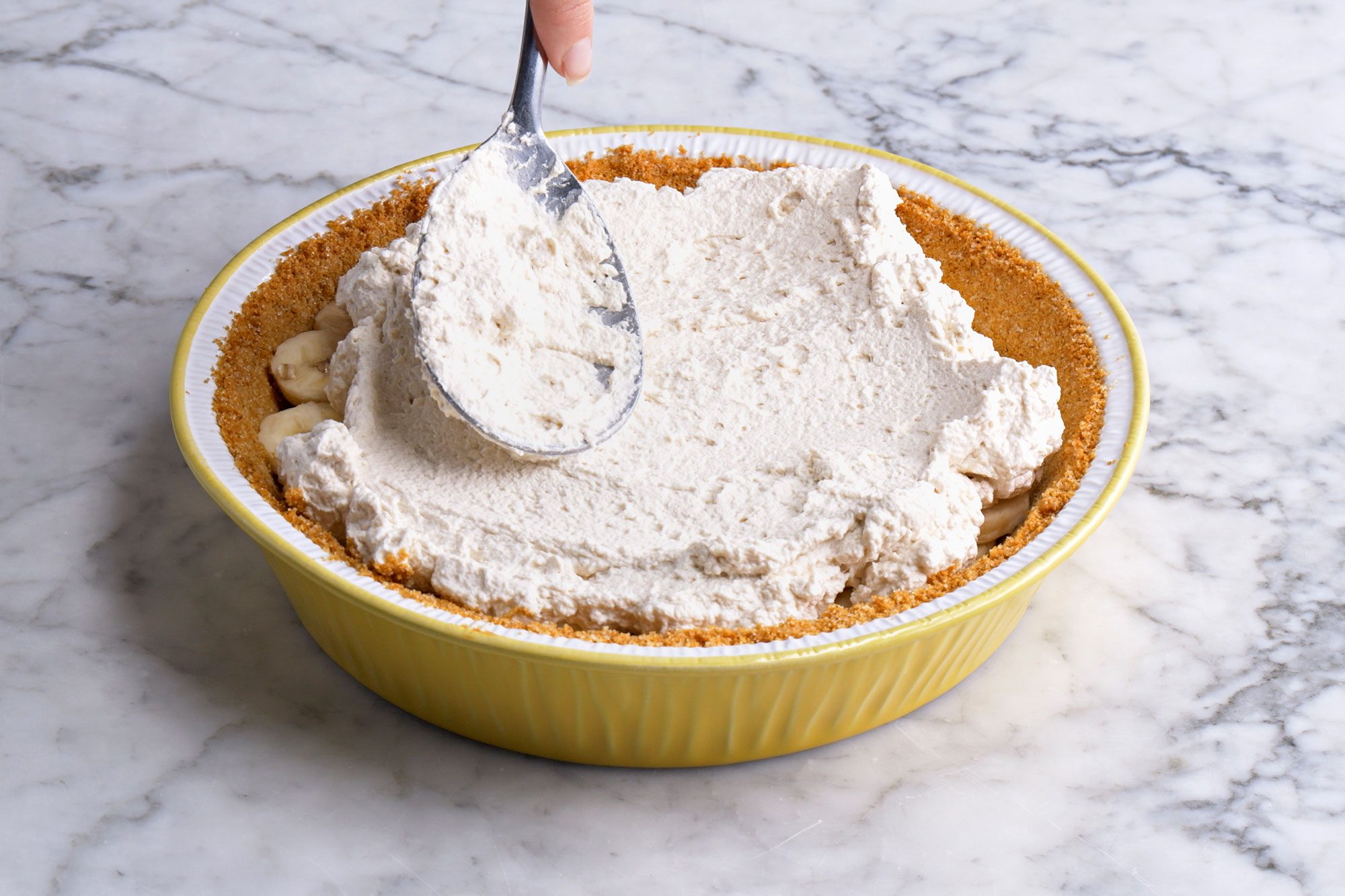 3/4 angle view shot of spoon whipped cream over the bananas; Refrigerate at least three hours or overnight; all set on a marble textured surface