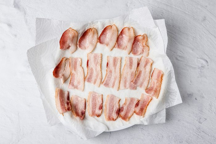 Cooked bacon strips arranged on a plate lined with white paper towels, set on a light gray surface. The paper towels are absorbing excess grease from the bacon.