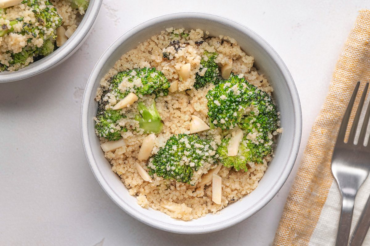 A white bowl filled with couscous, broccoli florets, and slivered almonds sits on a white surface next to a fork and a beige-striped napkin.