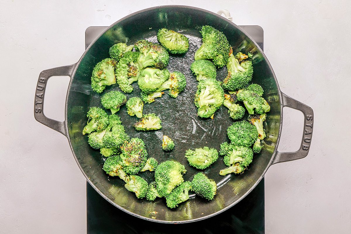A black pan on a stovetop contains several pieces of broccoli being sautéed, with some pieces showing slight browning.