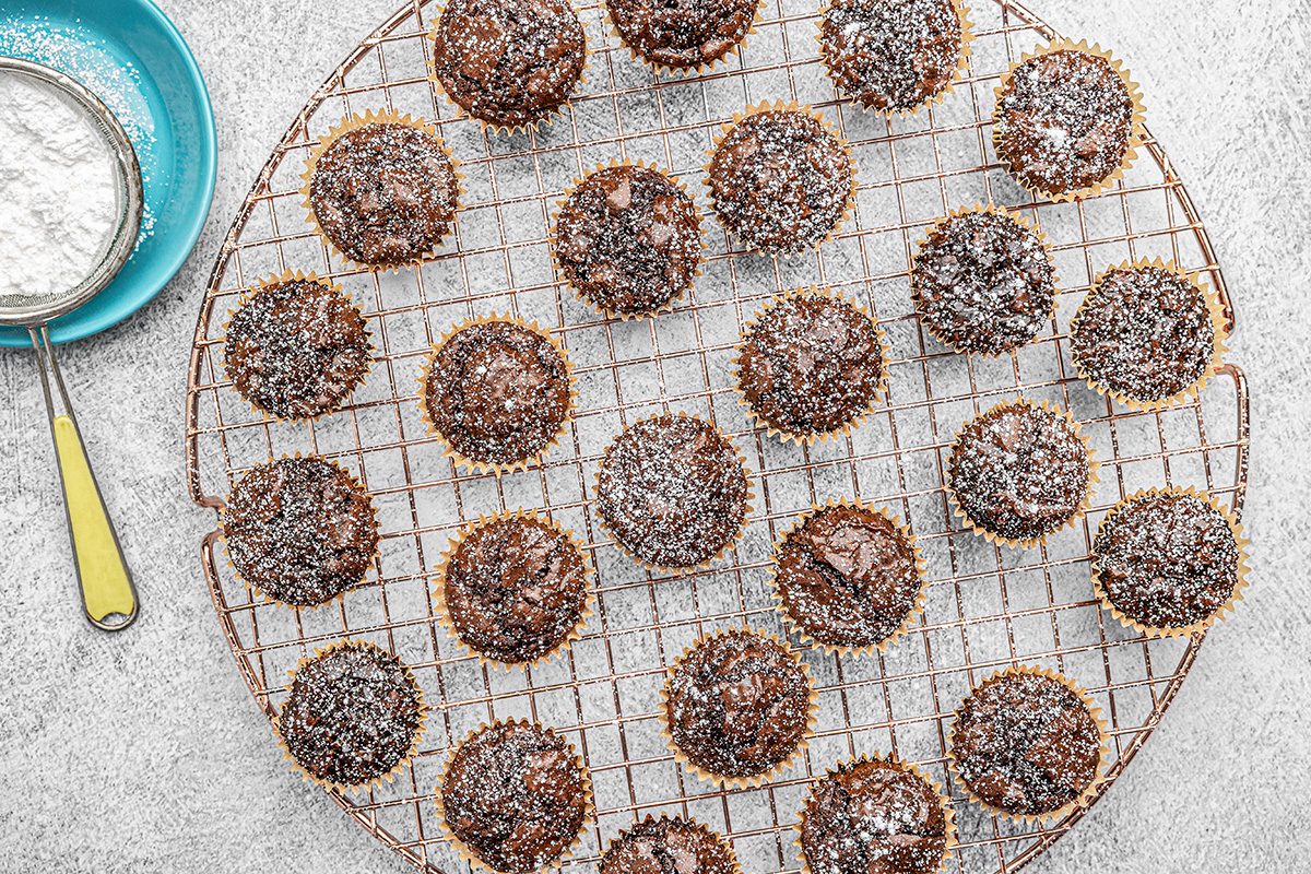 A round cooling rack holds several chocolate muffins dusted with powdered sugar. A small blue bowl and a fine mesh sieve with a yellow handle sit to the side on a light gray surface.