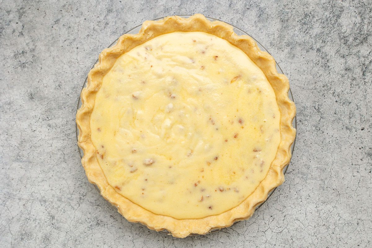 Unbaked quiche with a yellow egg mixture and bits of filling in a pie crust, set in a glass pie dish on a gray textured surface.