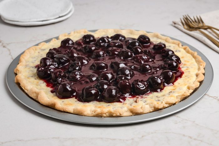 Close shot of Cherry Cheese Pizza on a pie plate; with ceramic plates and forks on a cream napkin arranged nearby; all set on a white texture marble surface