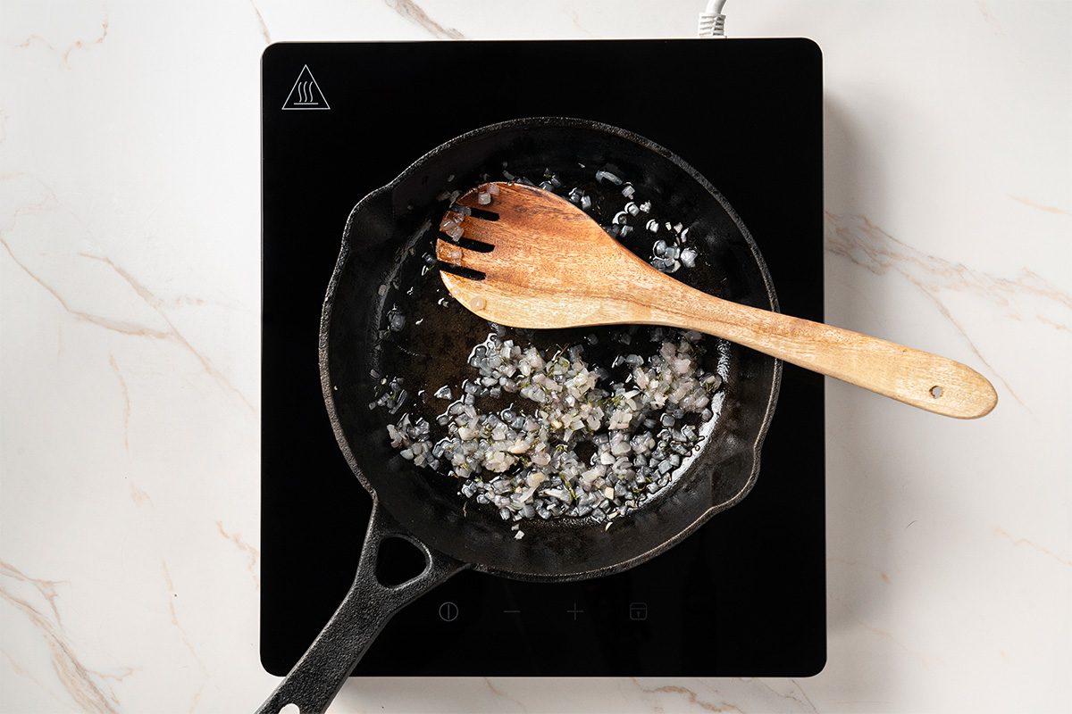 A cast iron skillet on an induction cooktop with chopped onions sautéing in oil, stirred by a wooden spoon on a white marble countertop.