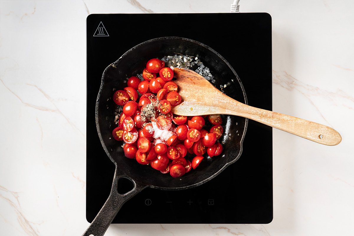 A cast iron skillet with halved cherry tomatoes and a wooden spoon sits on an electric stovetop, viewed from above.