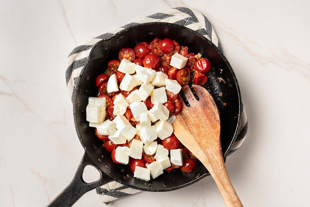 A cast iron skillet with halved cherry tomatoes and cubes of feta cheese, placed on a striped cloth. A wooden spatula rests inside the skillet on a light surface.
