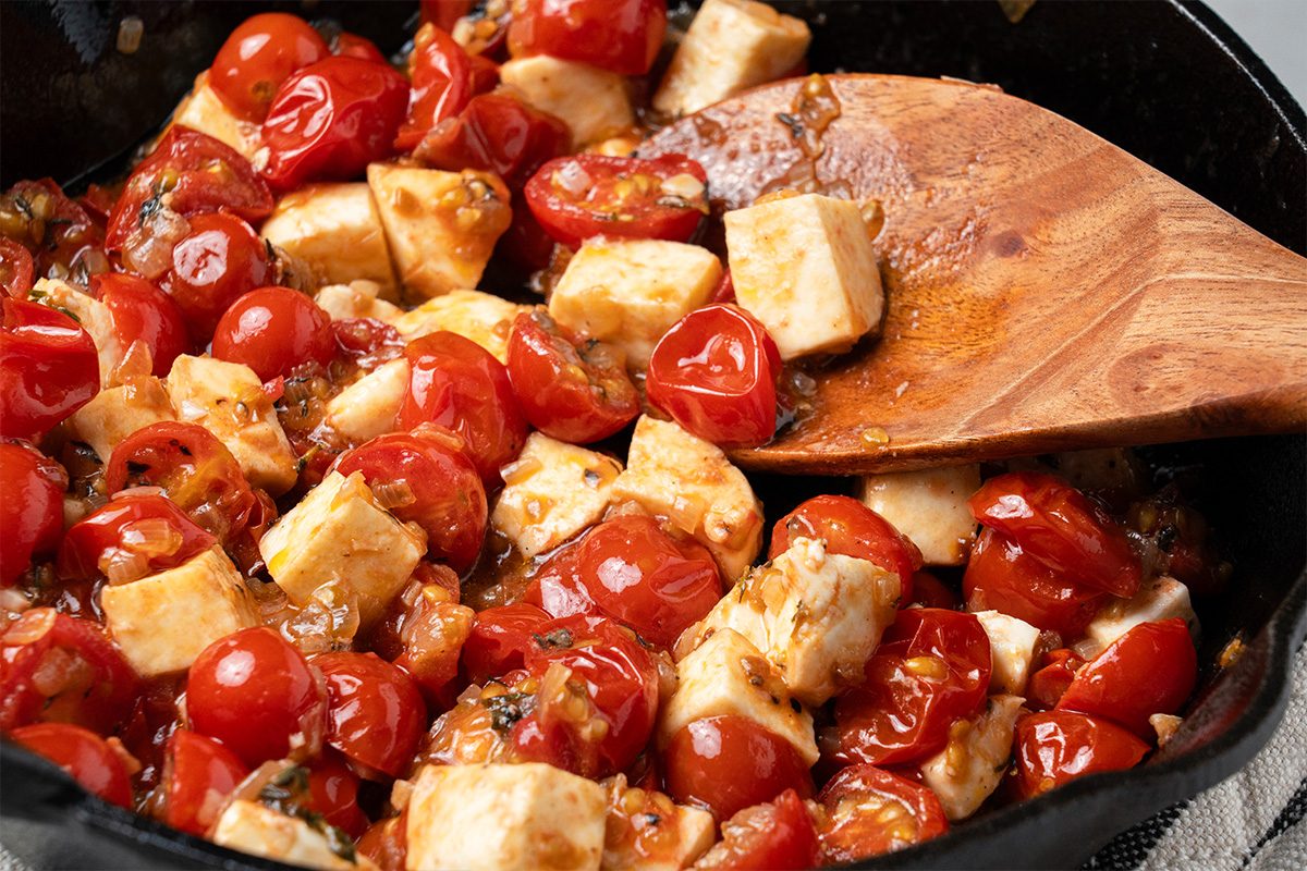 A close-up of a skillet filled with sautéed cherry tomatoes and cubes of tofu being stirred with a wooden spoon.