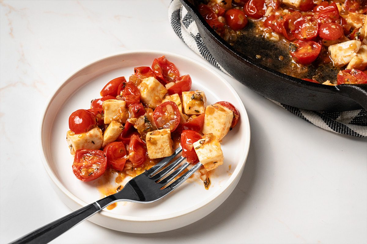 A white plate with a fork holds a serving of baked cherry tomatoes and cubes of mozzarella cheese, with a baking dish containing more of the same dish nearby on a white surface.