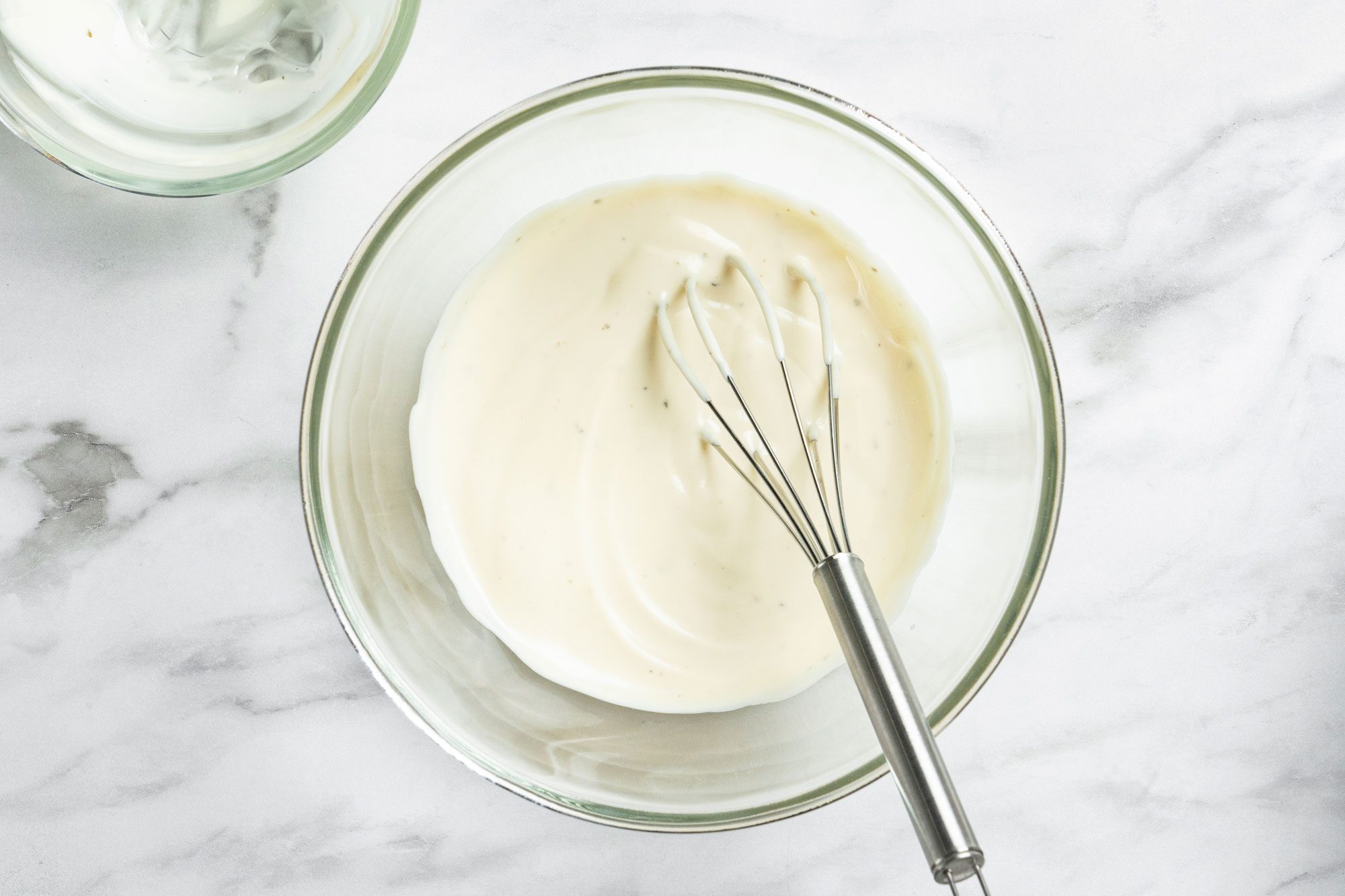 Overhead shot of a small bowl whisk the ranch dressing and mayonnaise; whisk tool; all set on a marble surface