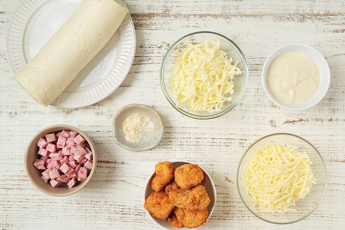 A flatlay of ingredients on a white wooden surface, including a tortilla wrap, two bowls of shredded cheese, a bowl of diced ham, breaded chicken pieces, grated Parmesan, and a bowl of white sauce.