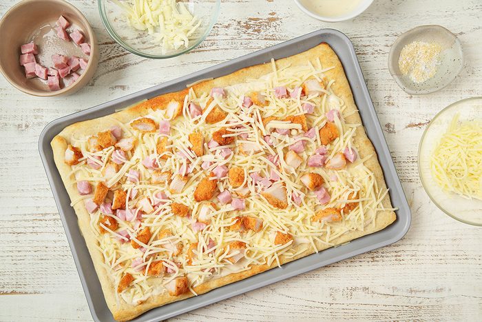 A rectangular pizza dough on a baking sheet topped with shredded cheese, diced ham, and chicken pieces. Surrounding the tray are bowls with more cheese, ham cubes, and sauce on a white wooden surface.
