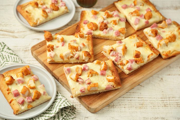 Rectangular slices of flatbread topped with melted cheese, diced ham, and chicken pieces are arranged on a wooden board. Two additional pieces are served on small round plates.