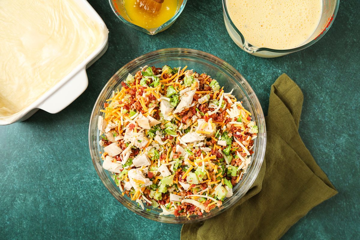 A glass bowl containing chopped vegetables; shredded cheese; and chicken is on a green surface beside a casserole dish with phyllo dough; a measuring cup of liquid; and a green cloth napkin. The background is simple and uncluttered