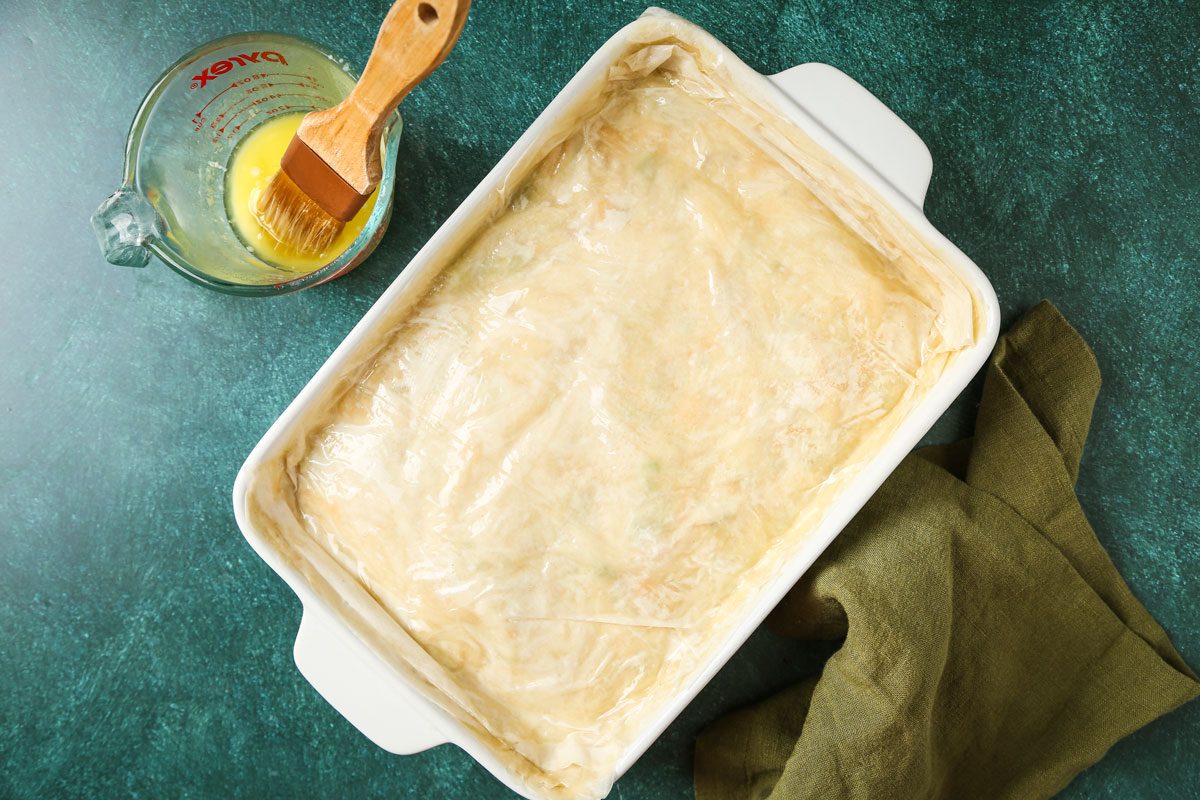 A rectangular baking dish layered with phyllo dough rests on a green surface; a measuring cup holding egg wash and a pastry brush sit beside it; a green cloth lies nearby