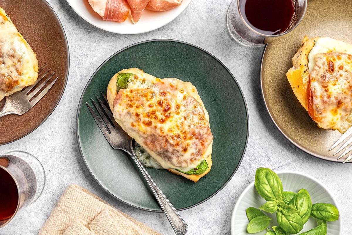 Three plates with cheesy baked chicken breasts, some with vegetables underneath, are arranged on a table with glasses of red wine, a bowl of cured meat, basil leaves, and a napkin. A fork rests on one plate.