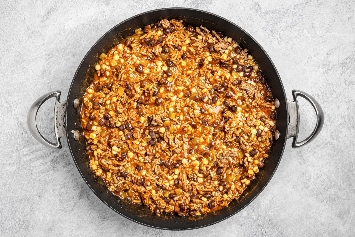 overhead shot of a large black skillet filled with a hearty mixture of ground meat, black beans, corn, diced tomatoes, and sauce, on a light gray surface