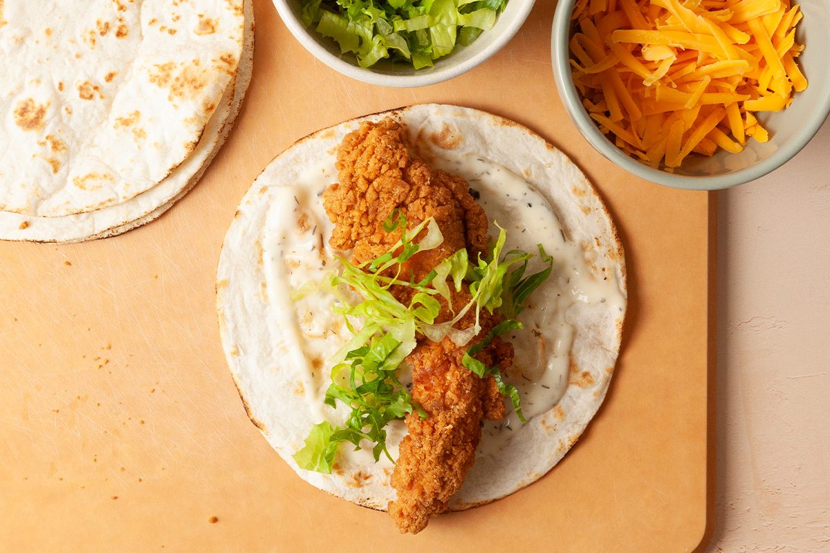 A flour tortilla topped with fried chicken, shredded lettuce, and shredded cheese sits on a cutting board, with bowls of lettuce and cheese nearby.